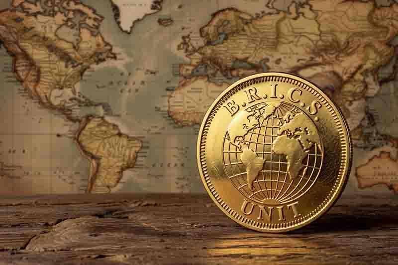 Eine große, goldglänzende Münze steht aufrecht im rechten Vordergrund auf einer rustikalen, dunklen Holzoberfläche. Die Münze zeigt in der Mitte einen Globus mit Längen- und Breitengraden; am oberen Rand ist der Schriftzug „B.R.I.C.S.“ und am unteren Rand das Wort „UNIT“ eingraviert.